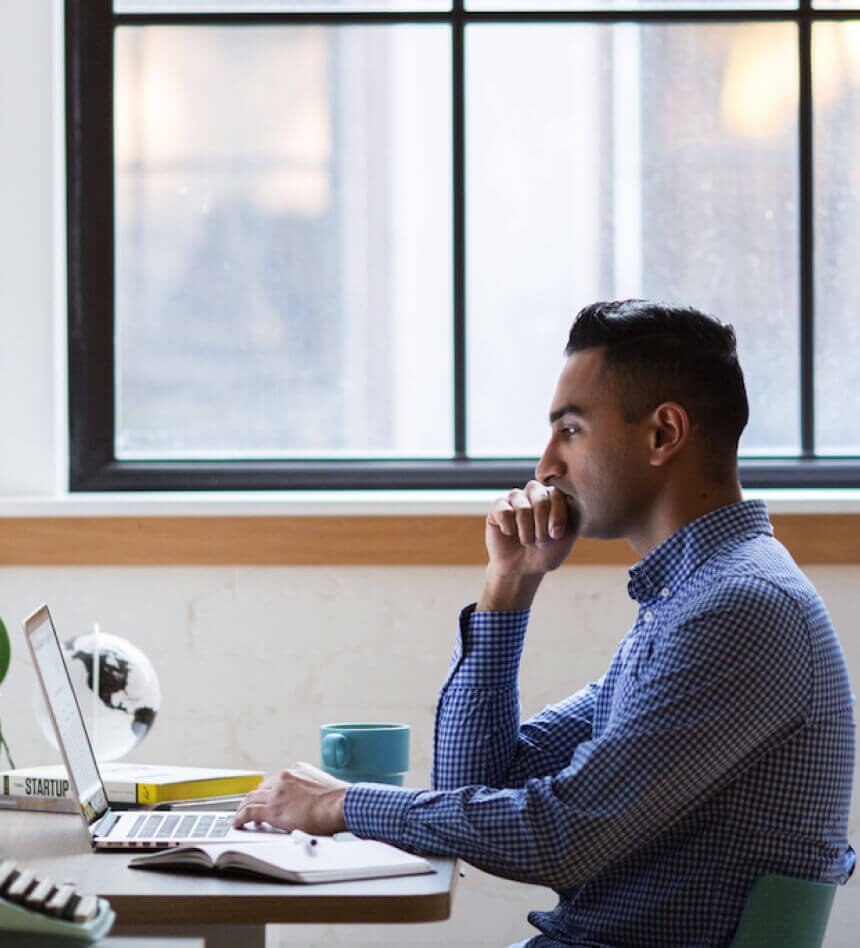 Man working on a laptop with a notebook and coffee mug by the window