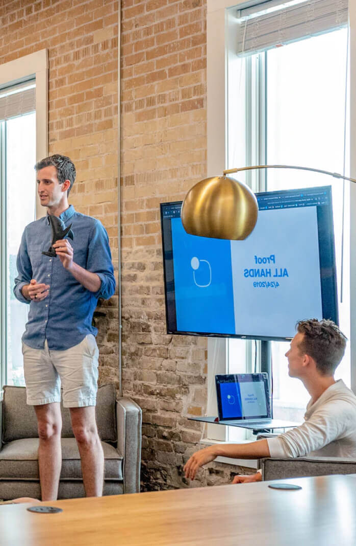 Man giving a presentation in an office with a screen displaying a slide and colleague listening
