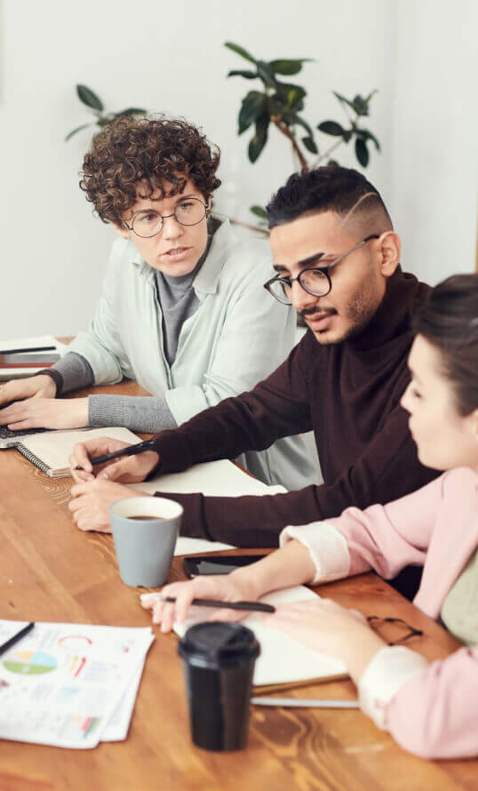 Three colleagues collaborating at a wooden desk with reports, notebooks, and coffee