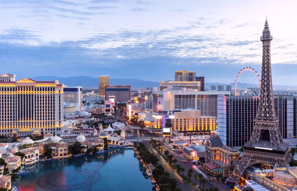 A panoramic view of the Las Vegas Strip at dusk, featuring famous landmarks such as the Eiffel Tower replica, Caesars Palace, and the High Roller observation wheel, with a reflective water fountain and distant mountains under a partly cloudy sky.