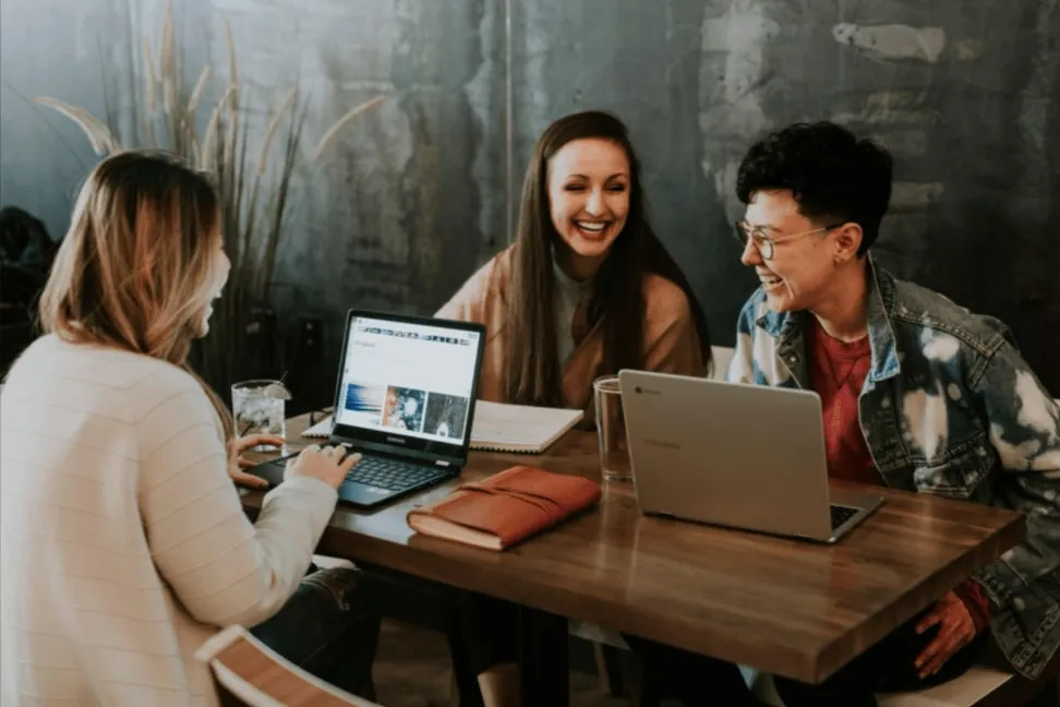 Three young professionals smiling and working together on laptops at a café-style workspace
