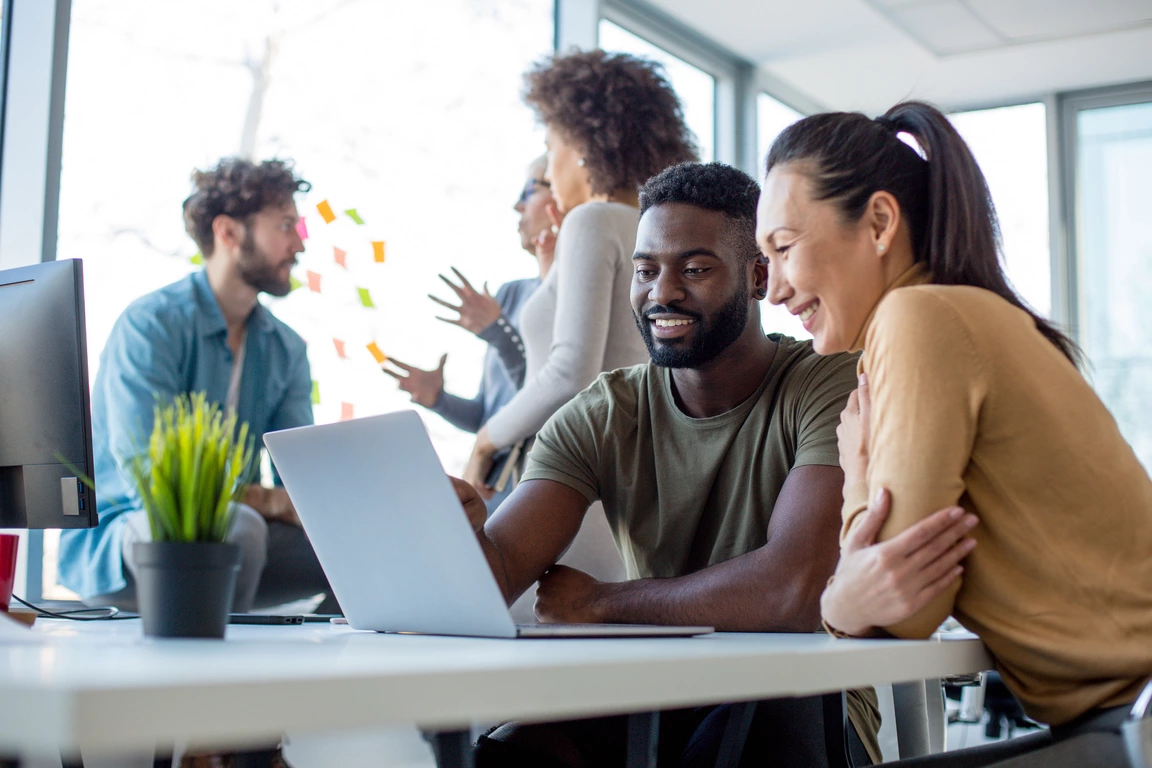 Two colleagues collaborating on a project using a laptop in a busy office environment, focusing on essential Local SEO strategies.