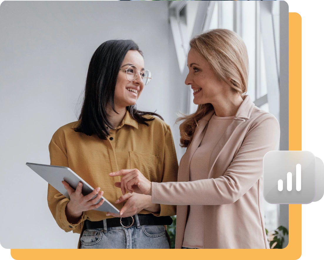 Two women stand indoors, one holding a tablet while the other points at the screen. Both are smiling and discussing SEO services. A chart icon is present in the lower right corner, hinting at their focus on website performance.