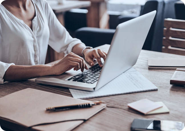 Person typing on a laptop at a wooden desk with papers, envelopes, a pen, sticky notes, and a smartphone—perhaps researching Local SEO Services. Blurred background with bookshelves and seating areas.