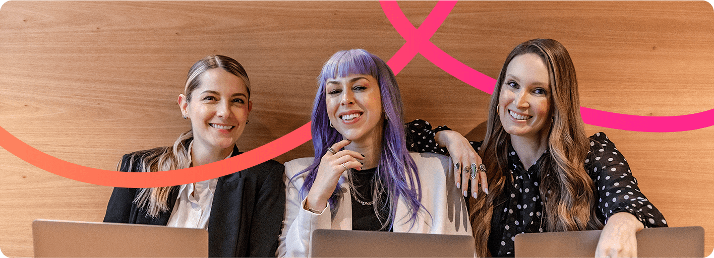 Three women sit together at a table with laptops, smiling, in front of a wooden wall with pink curved lines, collaborating on private label SEO strategies.