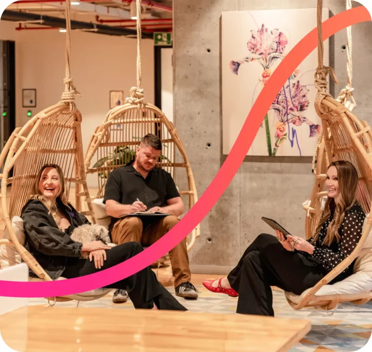 Three people sit on hanging wicker chairs in a modern indoor space; one holds a dog, another writes on a notepad, and the third uses a tablet.