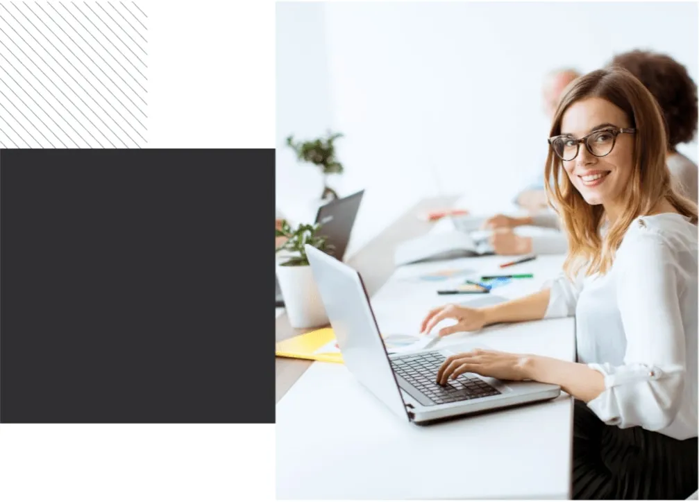 Professional woman smiling at the camera while working on a laptop in a modern office environment with colleagues in the background.