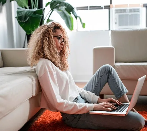 Young woman with curly hair sitting on the floor using a laptop in a cozy modern living room with natural light and houseplants.