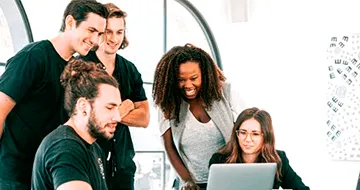 Five people gather around a desk, looking at a laptop screen and smiling in a bright, modern office setting, collaborating on digital PR services.