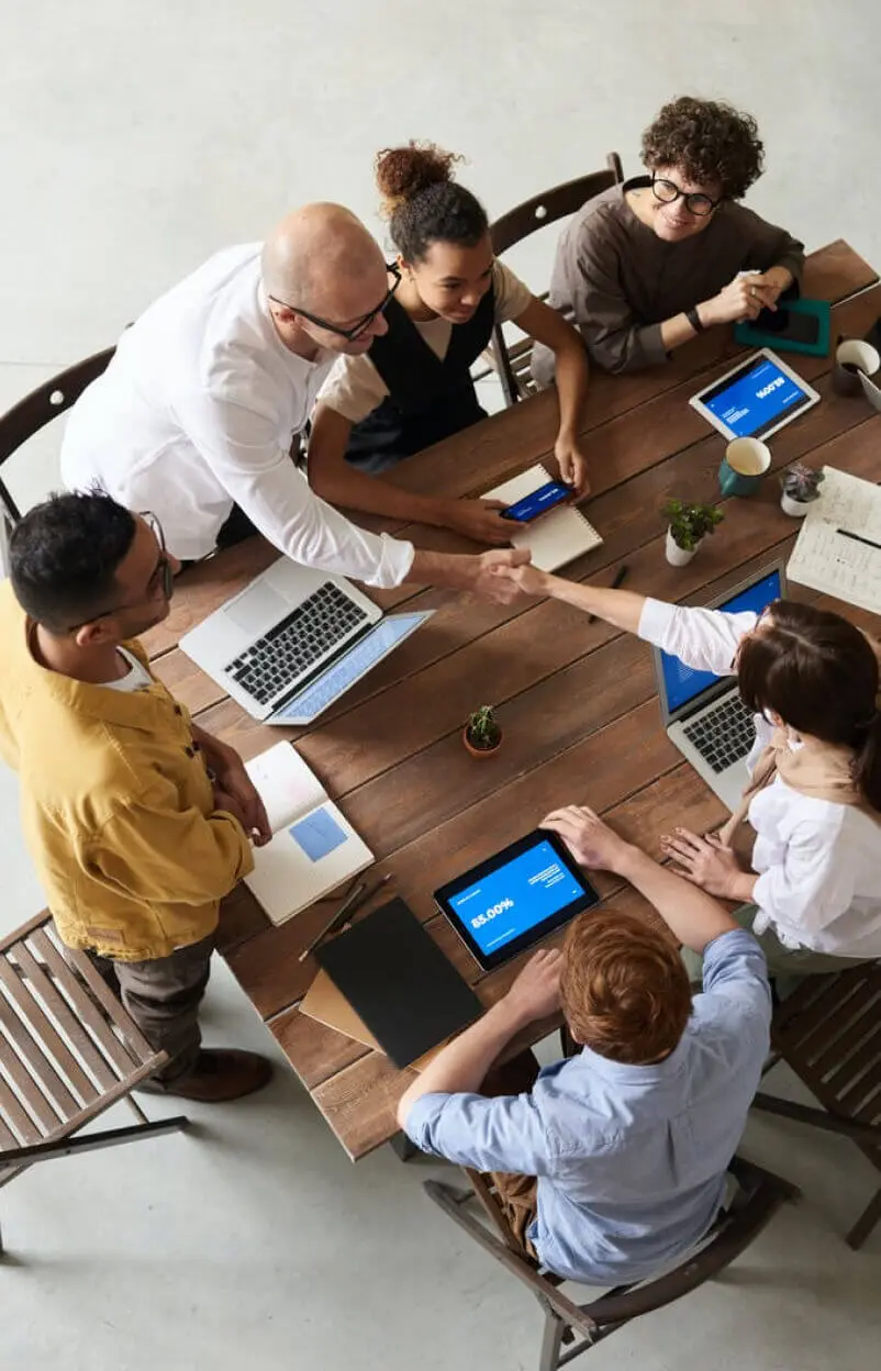 Team of professionals in a meeting around a wooden table shaking hands and discussing data on laptops and tablets